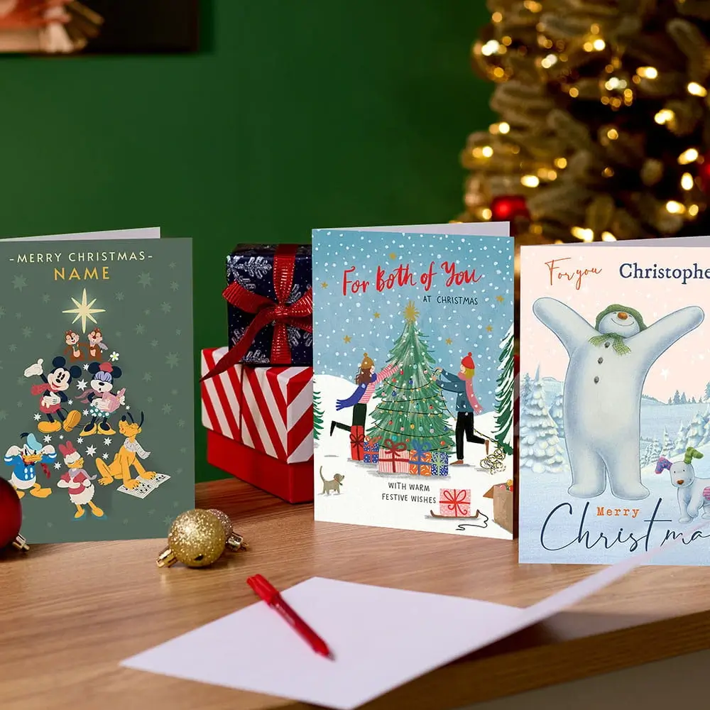 Selection of Christmas cards, including Disney, festive and personalised designs, displayed on a wooden table with wrapped presents and a decorated Christmas tree in the background. Selection of Christmas cards, including Disney, festive and personalised designs, displayed on a wooden table with wrapped presents and a decorated Christmas tree in the background.